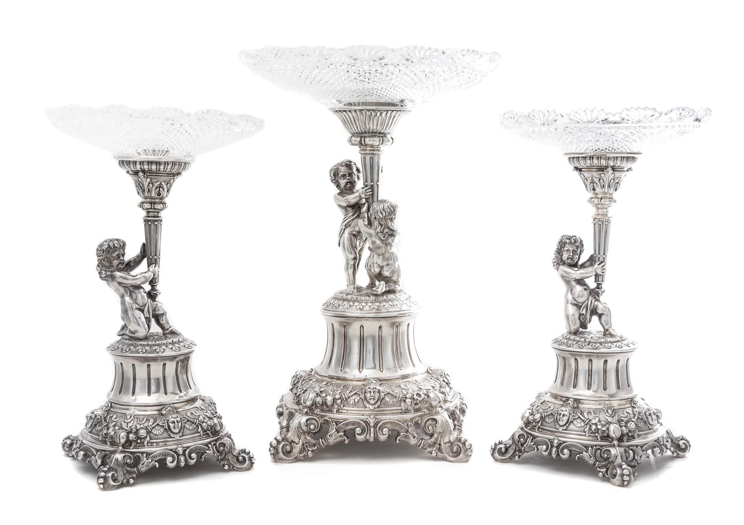 A German Silver and Wheel Cut Glass Three-Piece Figural Garniture, Late 19th Century, each standard modeled as two putti or a putto hol