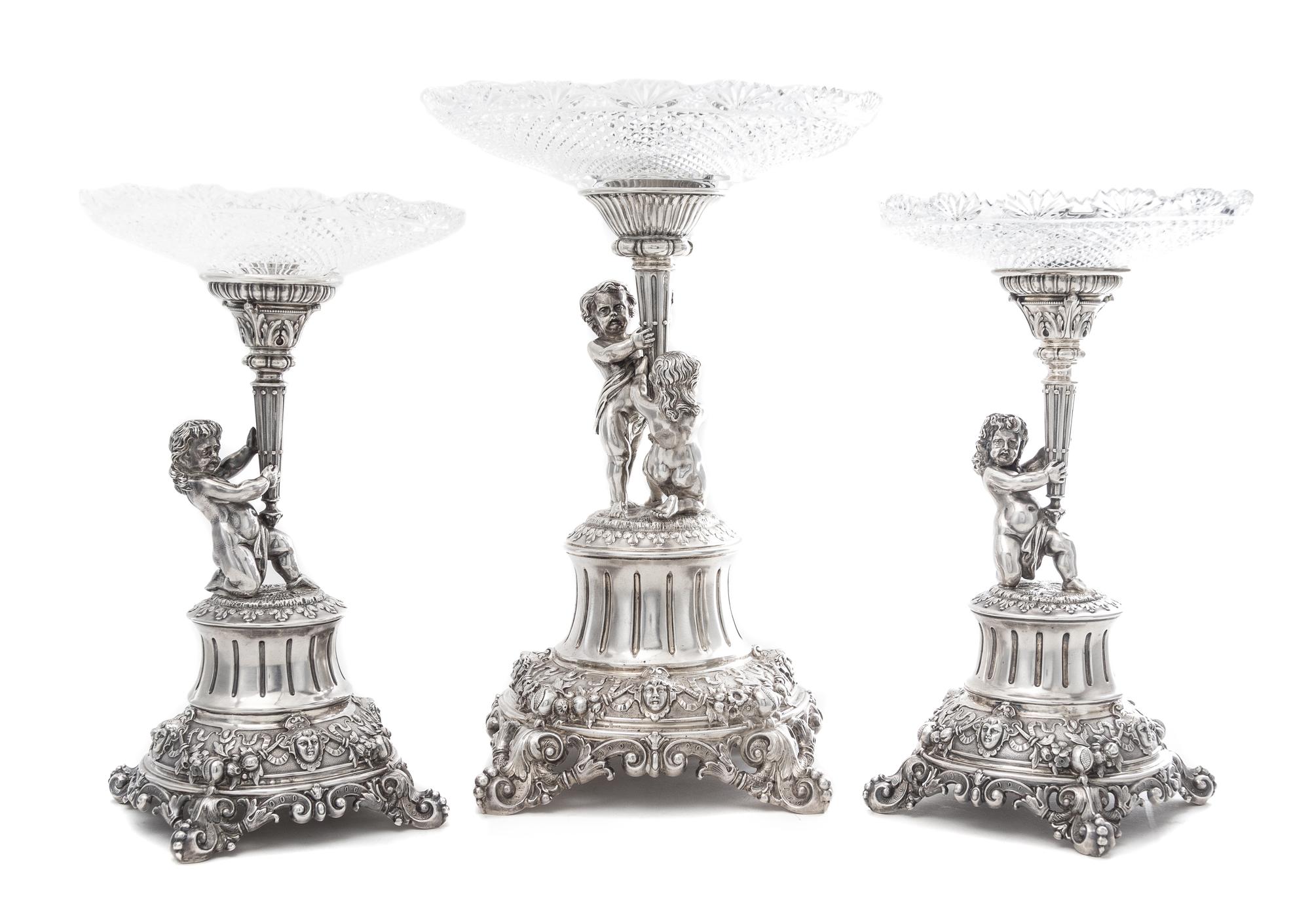 A German Silver and Wheel Cut Glass Three-Piece Figural Garniture, Late 19th Century, each standard modeled as two putti or a putto hol