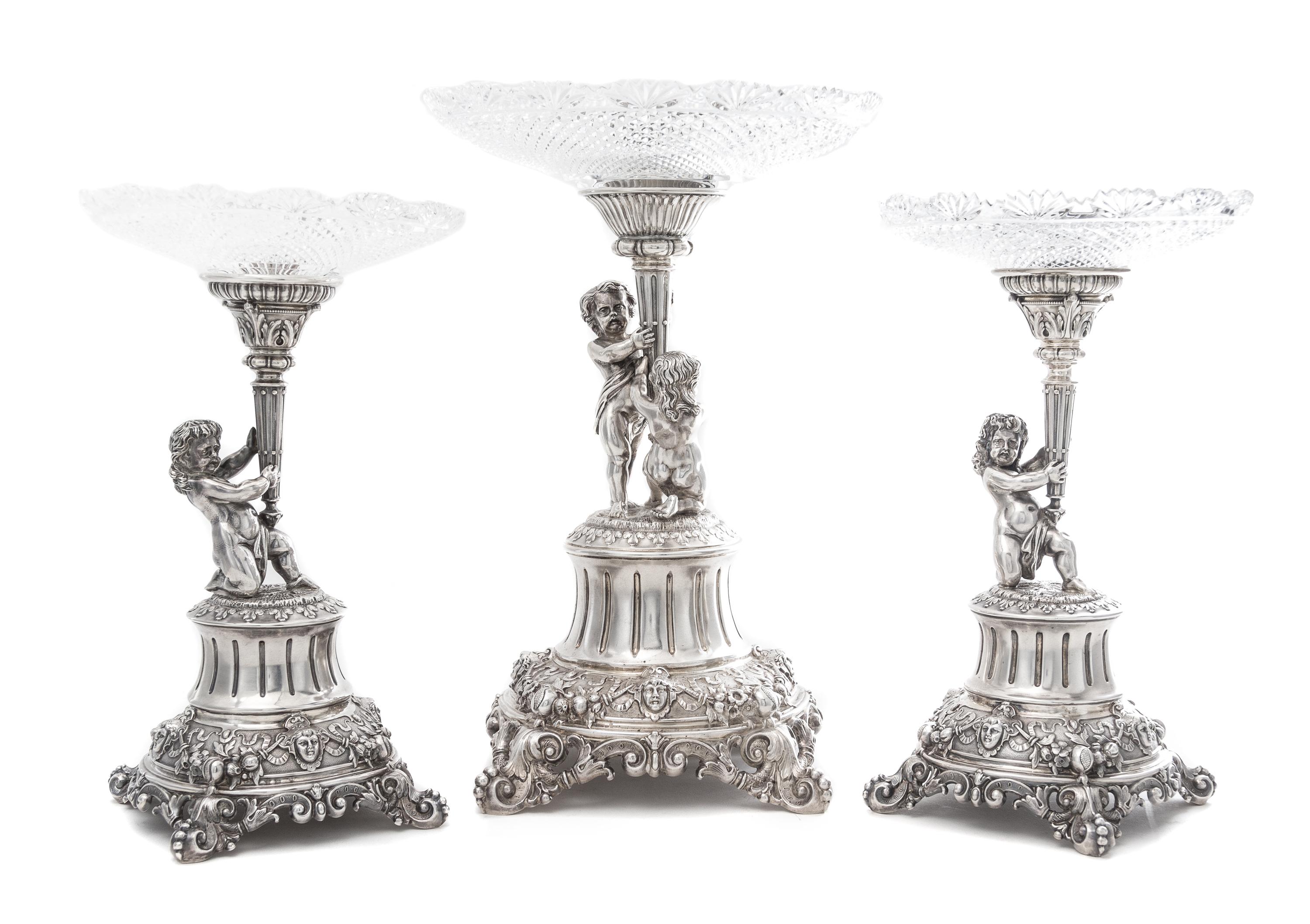 A German Silver and Wheel Cut Glass Three-Piece Figural Garniture, Late 19th Century, each standard modeled as two putti or a putto hol