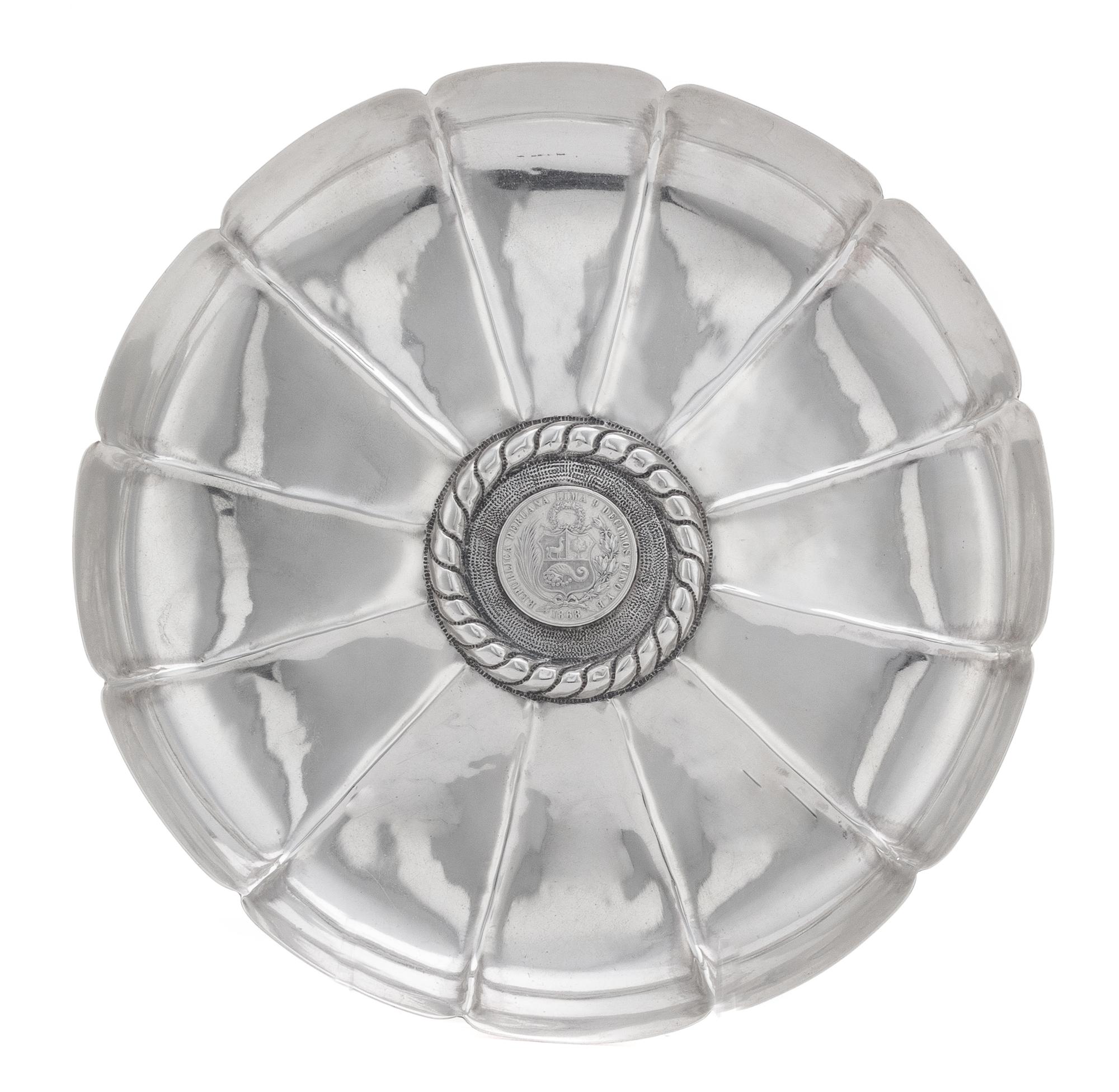 A Peruvian Silver Dish, , having a scalloped edge and a ribbed interior, centered by a banded motif surrounding an 1868 1 sol silver co