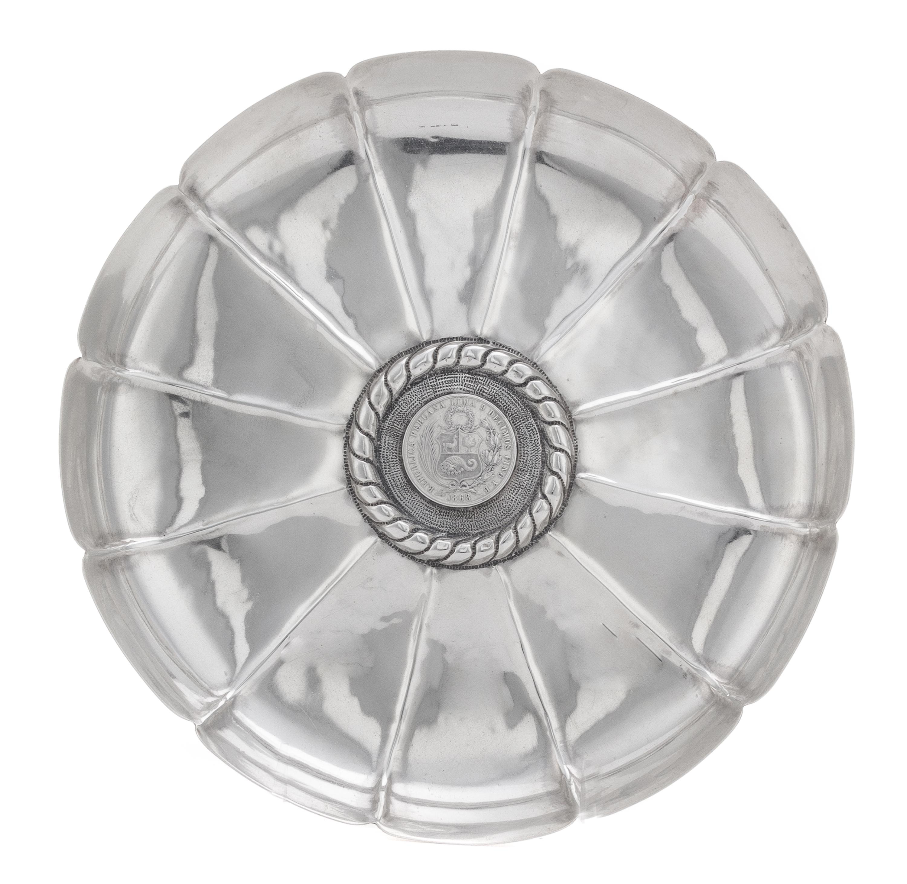 A Peruvian Silver Dish, , having a scalloped edge and a ribbed interior, centered by a banded motif surrounding an 1868 1 sol silver co