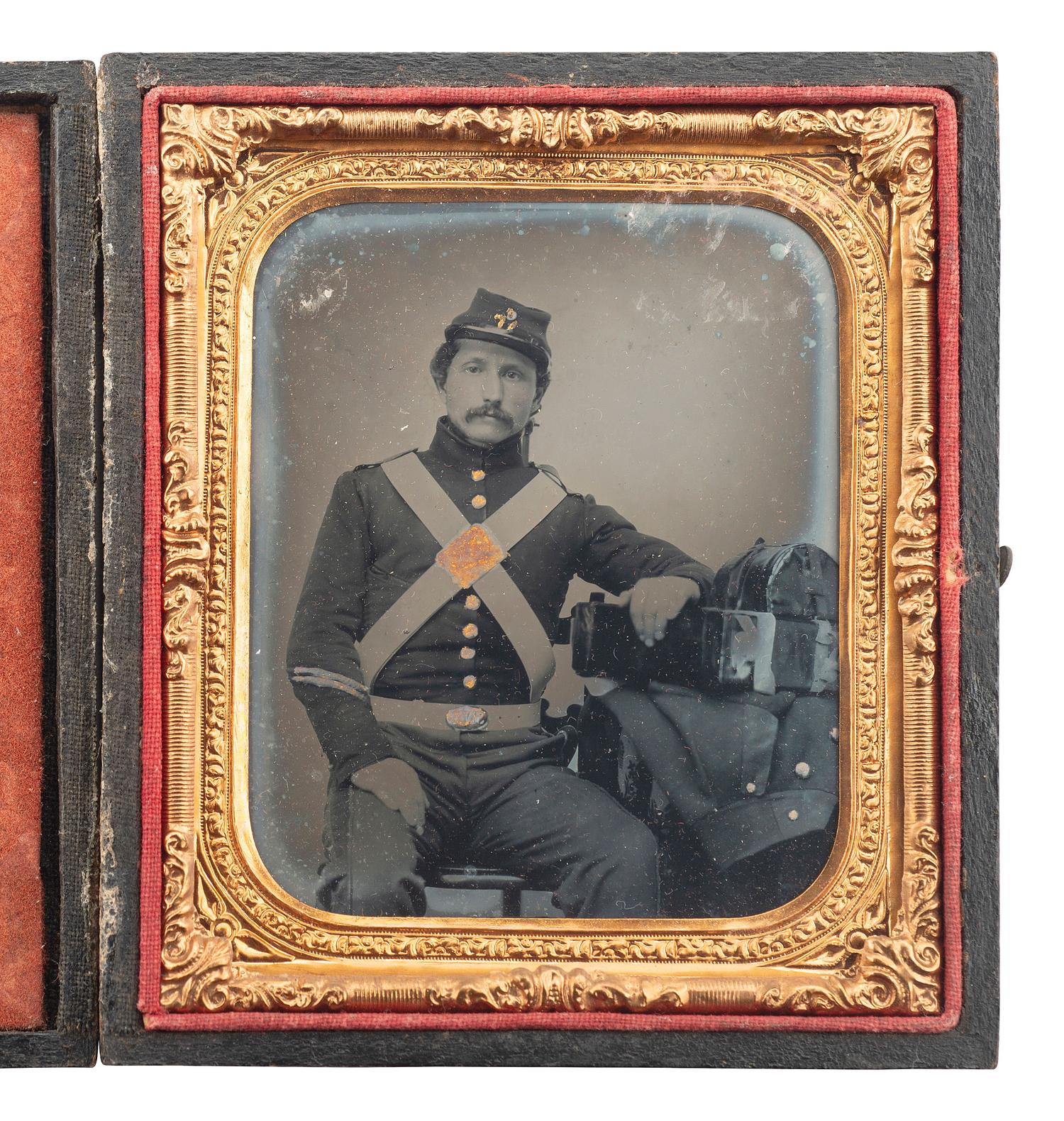 [CIVIL WAR]. Sixth plate ruby ambrotype portrait of a 71st New York State Militia private posed in uniform with his knapsack, rolled blanket, and great coat sitting on the table next to him.