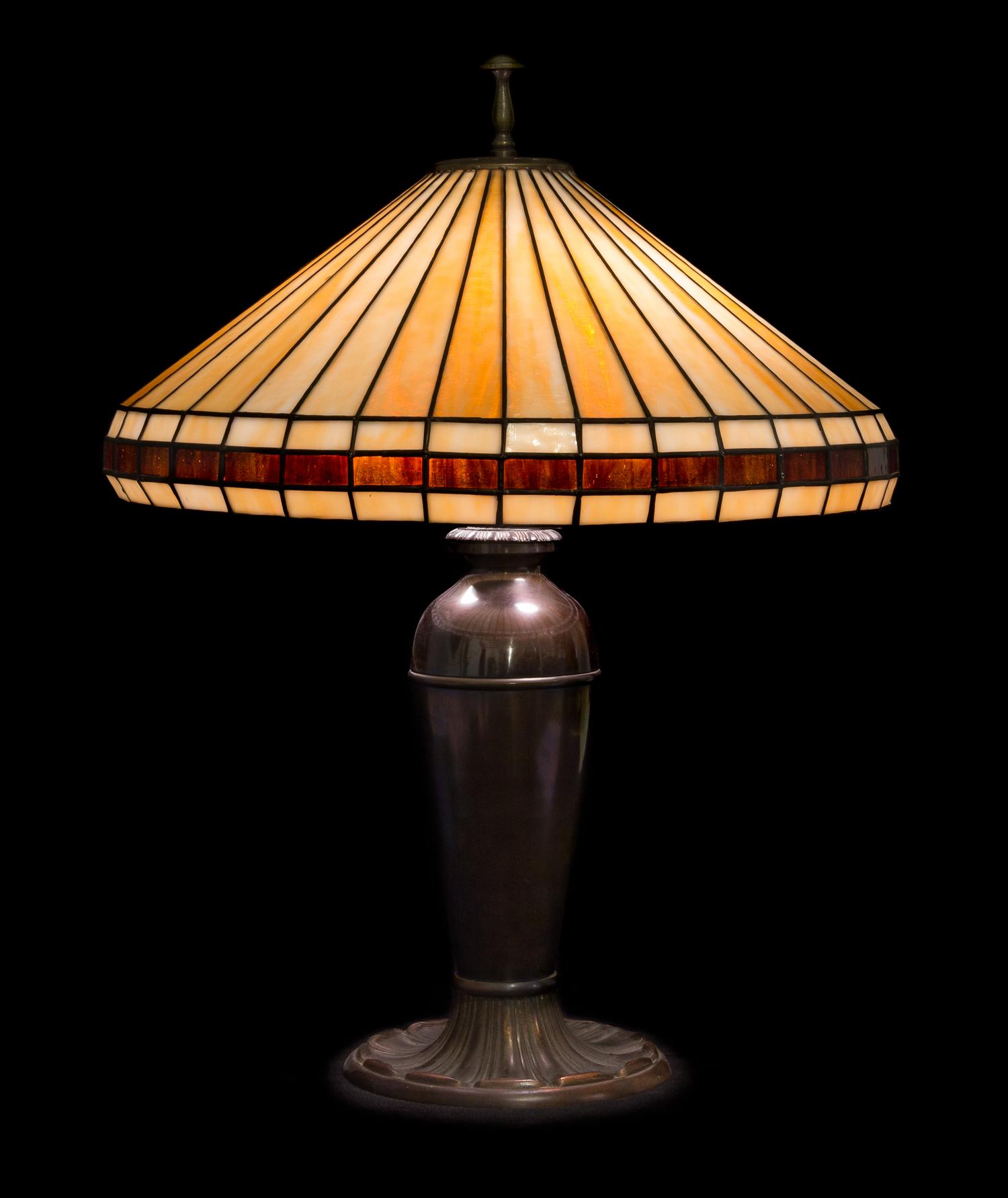 Miller Lamp Co., EARLY 20TH CENTURY, a leaded glass table lamp, having a conical geometric shade raised on an urn-form base